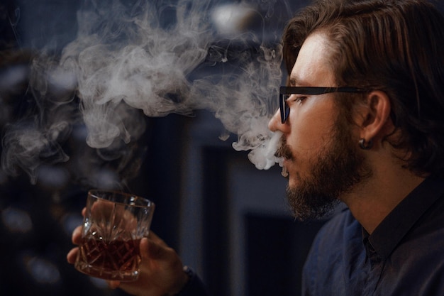 El hipster de pelo largo emite un espeso humo de cigarrillo. Hombre sosteniendo un vaso con alcohol