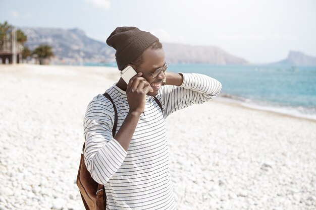 Hipster de piel oscura y despreocupada con ropa de moda conversando por teléfono inteligente mientras camina por la playa de guijarros y se relaja en el día de verano junto al mar