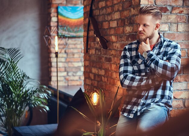 Un hipster rubio barbudo vestido con jeans y camisa polar posando en una habitación con interior de loft.