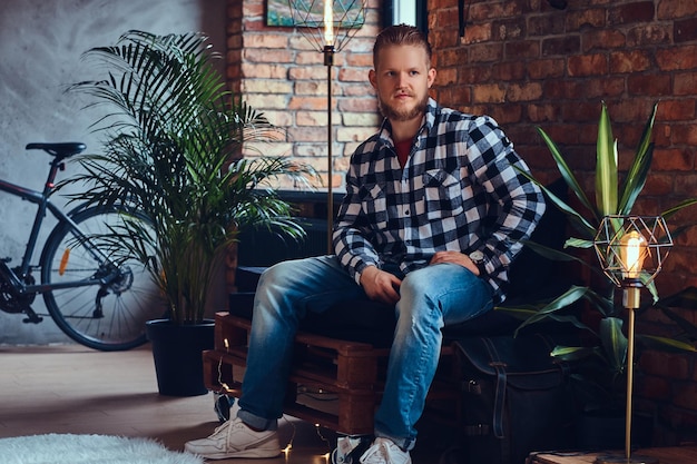 Un hipster rubio barbudo vestido con jeans y camisa polar posando en una habitación con interior de loft.