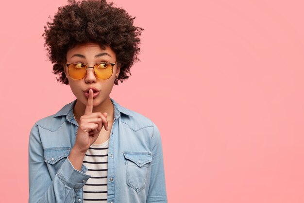 Hipster secreto de piel oscura quiere ocultar información, muestra un gesto de shh con el dedo índice sobre la boca, mira sospechosamente a un lado, vestido con ropa de moda, aislado sobre una pared rosa