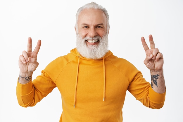 Hipster senior feliz con tatuajes que muestran el signo v de la paz y sonriendo, riendo feliz como vistiendo una sudadera con capucha moderna naranja y divirtiéndose, de pie sobre una pared blanca