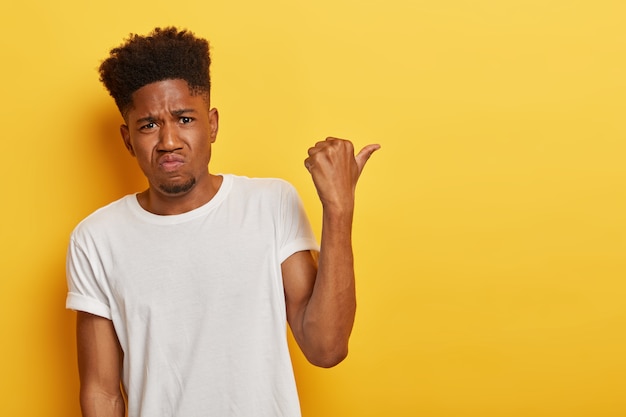 Hipster sombrío con piel oscura y cabello afro, apunta a un lado de mala gana, tiene expresión triste