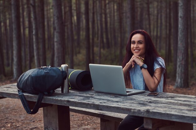 Hipster sonriente mirando a la cámara mientras se sienta en un banco de madera con un portátil abierto en un hermoso bosque de otoño.