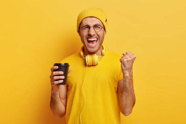 Hipster triunfante lleno de alegría aprieta el puño, exclama con satisfacción, sostiene café para llevar, tiene auriculares en el cuello