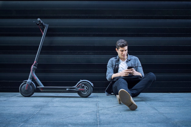 Hipster usando un teléfono inteligente mientras está sentado en el suelo junto a su scooter eléctrico contra la pared negra