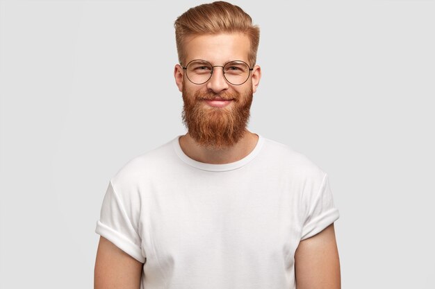 Hispster de moda alegre encantado con barba pelirroja, lleva gafas redondas y camiseta blanca