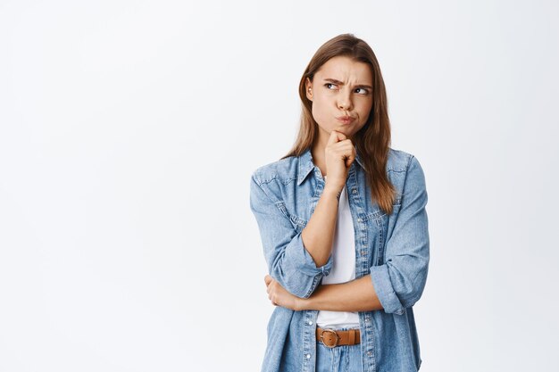 Hmm, algo extraño. Mujer joven pensativa que tiene dudas, frunciendo el ceño y haciendo pucheros mientras piensa, mirando la esquina superior izquierda pensativa, de pie sobre blanco