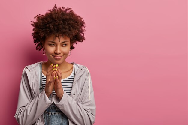 Hmm, buen plan. Mujer astuta y rizada mete los dedos, mira misteriosamente a un lado, tiene pensamientos o intenciones engañosas, quiere hacer algo malo, usa anorak, se para contra la pared rosa pastel, espacio libre