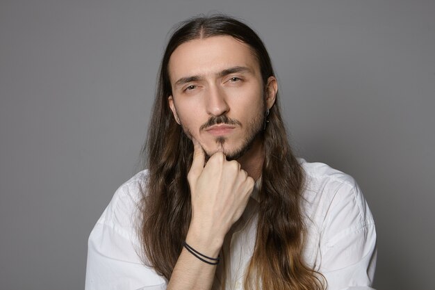 Hmm. Déjame pensar. Disparo aislado de serio concentrado joven elegante barbudo hombre de pelo largo en camisa blanca formal estrechando los ojos y sosteniendo la mano en la barbilla, con expresión facial sospechosa