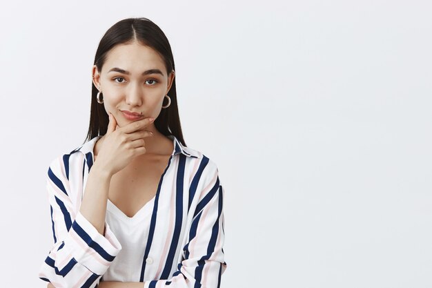 Hmm, déjame pensar. Retrato de mujer de moda inteligente y creativa pensativa con cabello largo y oscuro, frotándose la barbilla y sonriendo mientras hace un plan o una elección, decidiendo algo