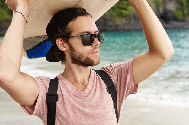 Hobby y vacaciones. Apuesto joven con barba con elegantes gafas de sol y snapback sosteniendo la tabla de surf sobre su cabeza, mirando hacia el océano, esperando grandes olas