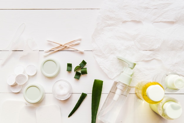 Hoja de papel mascarilla; cotonetes; Crema hidratante y aloevera sobre tabla de madera blanca.