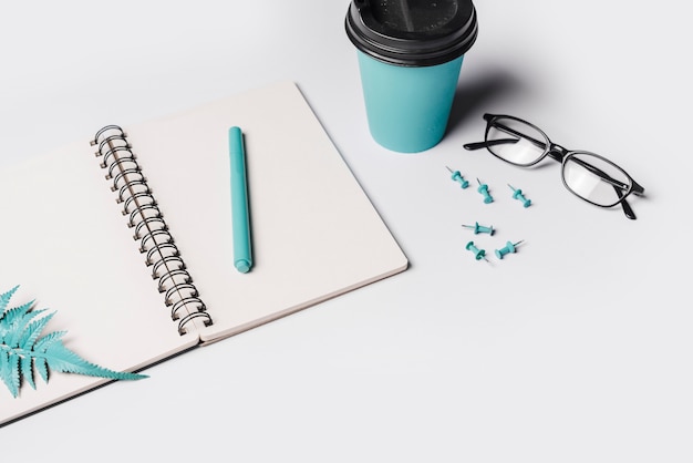 Hojas y pluma artificiales del helecho en el cuaderno espiral en blanco blanco con la taza de café; anteojos y alfiler