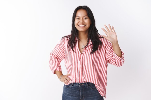 Hola amigos, ¿qué pasa? Amable y sociable linda encantadora mujer asiática en blusa a rayas saluda a los novatos agitando la mano con alegría en gesto de hola o hola sonriendo ampliamente a la cámara sobre la pared blanca