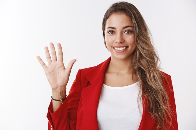 Hola. Chica saludando diciendo hola sonriendo amistosamente mirando positivo levantando la palma saludo, dando la bienvenida a los novatos que ingresan a la compañía, de pie confiado y sonriendo agradablemente en la pared blanca