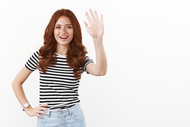 Hola, ¿cómo estás? Alegre, linda y linda adolescente pelirroja con camiseta a rayas levantando la mano y saludando felizmente sonriendo diciendo hola bienvenidos invitados saludando a los visitantes alegremente de fondo blanco