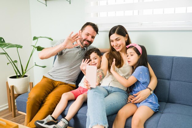 ¡Hola familia! Hermosa familia de cuatro miembros saludando mientras saluda a sus amigos y seres queridos durante una videollamada en línea en el teléfono inteligente