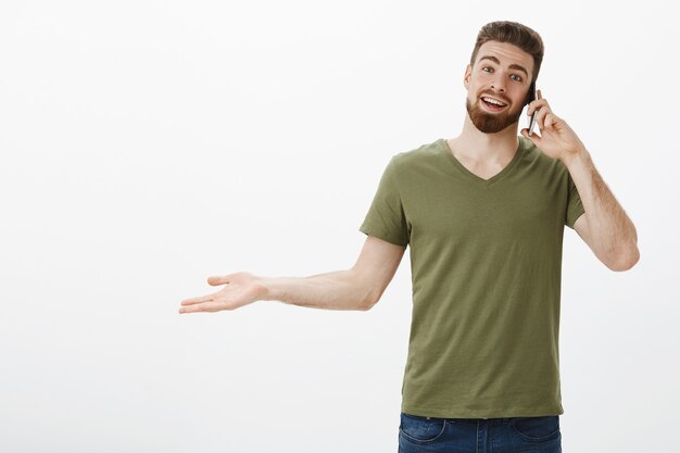 Hola, feliz recibe la llamada de tu amigo. Retrato de hombre barbudo atractivo joven complacido hablando por teléfono inteligente y extender la mano hacia los lados como sorprendido y contento de charlar contra la pared blanca