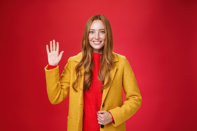 Hola, gusto en conocerte amigo. Mujer de aspecto amistoso femenino y elegante joven pelirroja linda en abrigo amarillo cálido otoño agitando la mano levantada en gesto de saludo y hola sonriendo ampliamente sobre la pared roja.