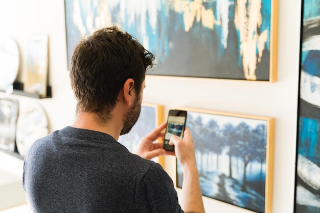 Hombre de unos 30 años interesado en comprar una obra de arte está tomando una foto de una pintura con su teléfono inteligente para mostrársela a su pareja