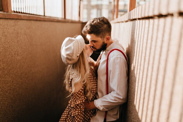 El hombre abraza a su novia rubia con gorra beige. Pareja romántica disfrutando de un día soleado y besos.