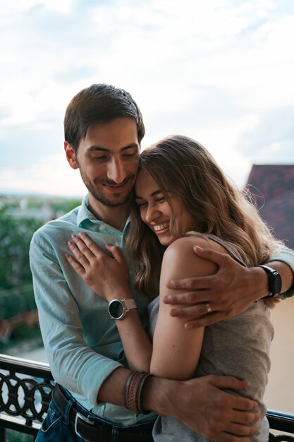 Hombre abrazando a esposa en el balcón. Pareja relajada disfruta de día y buenas noticias. Familia joven feliz