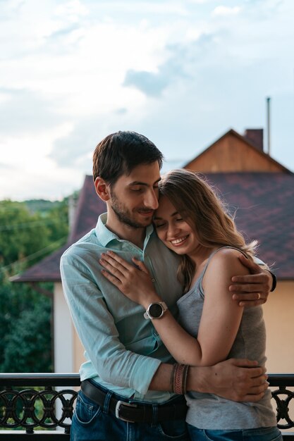 Hombre abrazando a esposa en el balcón. Pareja relajada disfruta de día y buenas noticias. Familia joven feliz