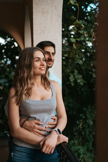 Hombre abrazando a esposa en el balcón. Pareja relajada disfruta de día y buenas noticias. Familia joven feliz