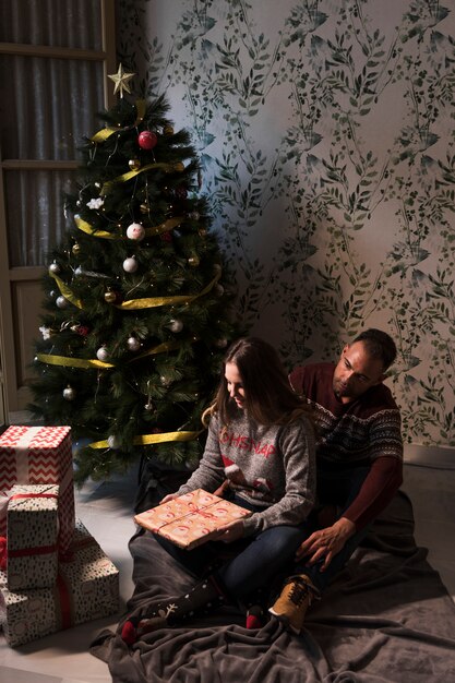 Hombre abrazando a una mujer desde atrás con un regalo en una colcha cerca del árbol de Navidad