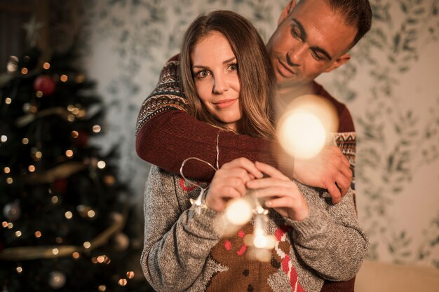 Hombre abrazando a la mujer desde atrás en suéteres cerca del árbol de Navidad
