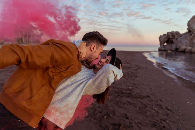 Hombre abrazando a mujer con bomba de humo rosa en la orilla del mar