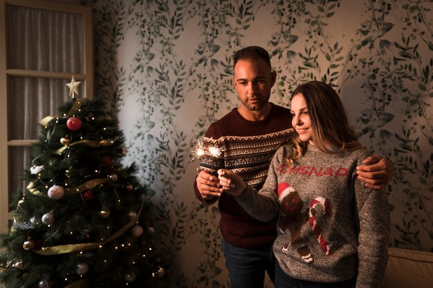 Hombre abrazando a mujer con luces de Bengala cerca de árbol de Navidad