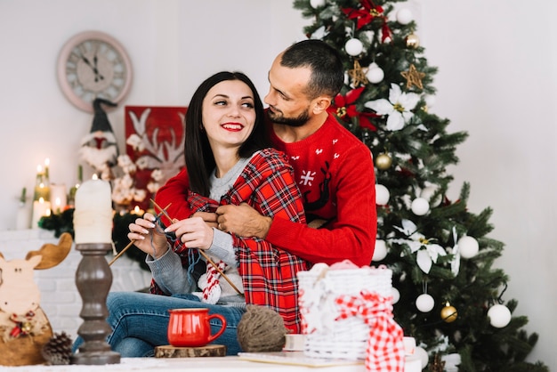 Hombre abrazando a mujer tejiendo cerca de árbol de Navidad