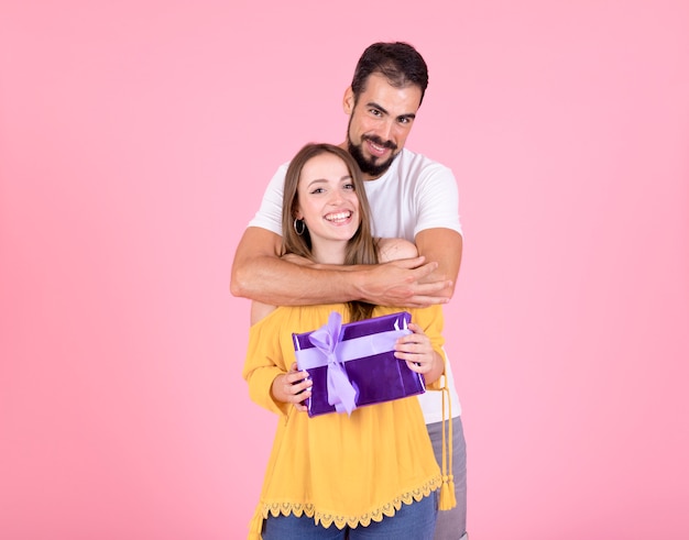 Hombre abrazando a su novia con caja de regalo púrpura sobre fondo rosa