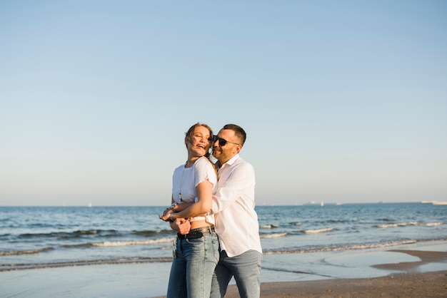 Hombre abrazando a su novia por detrás, de pie cerca del mar contra el cielo azul en la playa