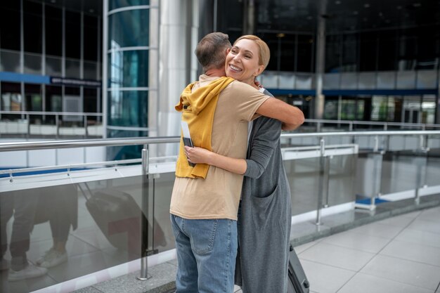 Hombre abrazando a una turista caucásica de mediana edad rubia y complacida sonriente en la terminal del aeropuerto