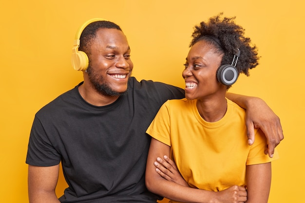 Hombre abrazar y mirar con amplias sonrisas el uno al otro escuchar música a través de auriculares vestidos con camisetas casuales aisladas en amarillo vivo