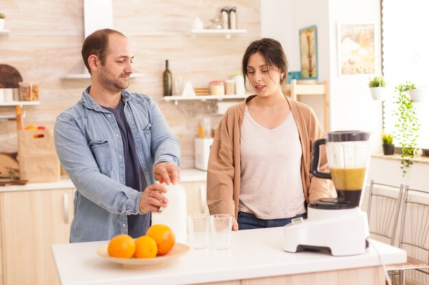 El hombre abre la botella de leche para batidos nutritivos mientras habla con su novia. Estilo de vida saludable, despreocupado y alegre, comiendo dieta y preparando el desayuno en una acogedora mañana soleada