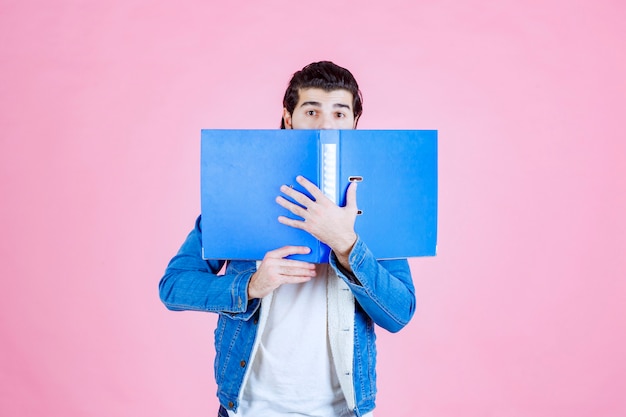 Hombre abriendo una carpeta azul y ocultando su rostro detrás de ella