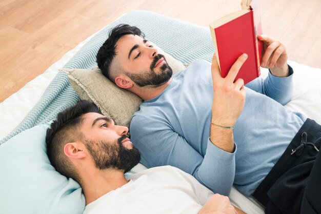 Hombre acostado en la acogedora cama mirando a su novio leyendo el libro