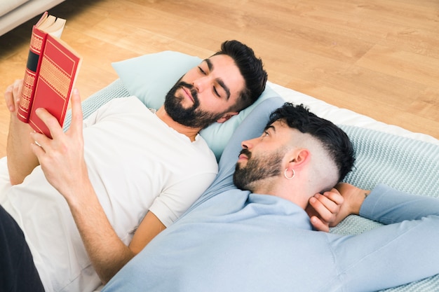 Hombre acostado en la cama mirando a su novio leyendo el libro