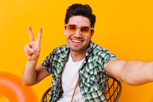 Hombre activo en traje a cuadros y gafas muestra el signo de la paz y hace selfie en espacio aislado.