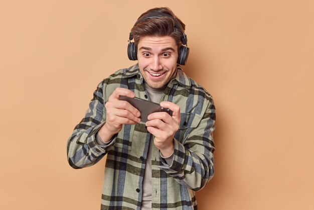 El hombre adicto juega videojuegos sostiene el teléfono inteligente horizontalmente trata de pasar un nivel difícil usa audífonos estéreo en las orejas vestido con una camisa a cuadros aislada sobre fondo beige Tecnología