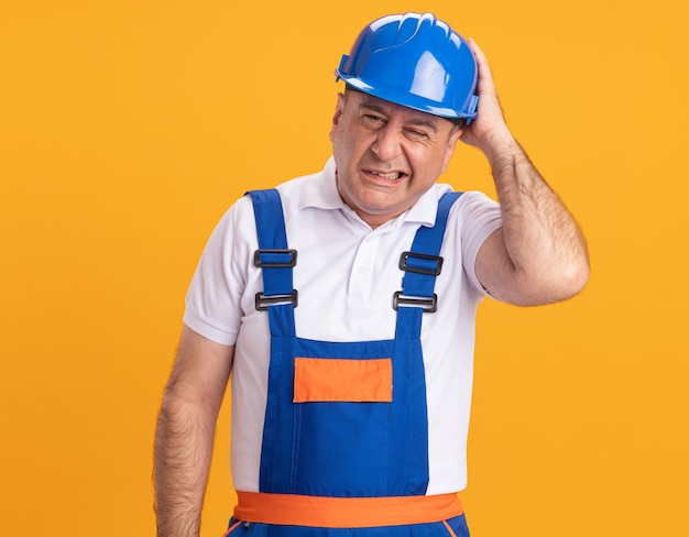 Hombre adulto adolorido constructor en uniforme sostiene la cabeza aislada en la pared naranja