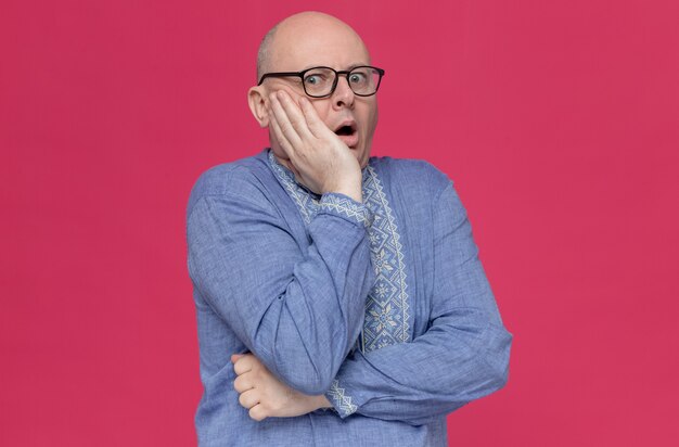 Hombre adulto ansioso en camisa azul con gafas poniendo la mano sobre su rostro y mirando