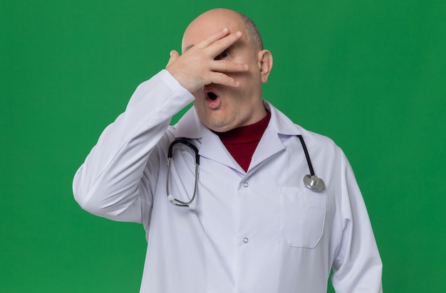 Hombre adulto ansioso en uniforme médico con estetoscopio poniendo la mano en la cara y mirando a través de los dedos