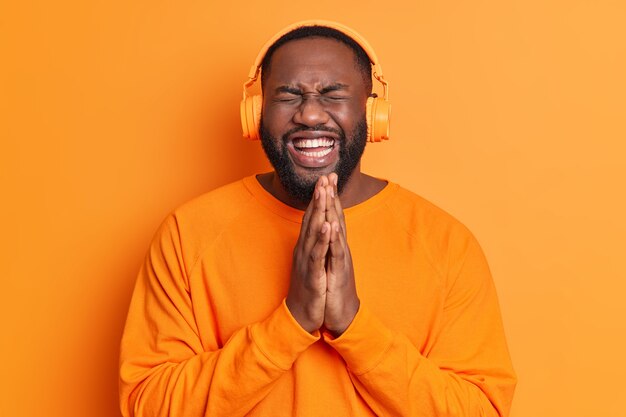 El hombre adulto con barba negra lleno de alegría mantiene las palmas presionadas juntas tiene un estado de ánimo optimista se ríe de algo gracioso usa auriculares estéreo vestidos con un suéter naranja aislado sobre una pared brillante