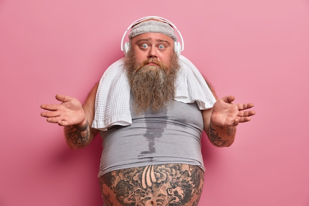 Un hombre adulto con barba y vacilante extiende las palmas de las manos y parece confundido, hace ejercicio regularmente para bajar de peso, escucha música en auriculares, usa una diadema y una camiseta de tamaño insuficiente, el vientre tatuado sobresale