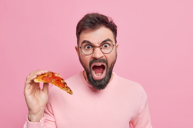 Hombre adulto barbudo emocional grita en voz alta sostiene una rebanada de sabrosa pizza apetitosa come comida rápida para bocadillos vestido con ropa casual